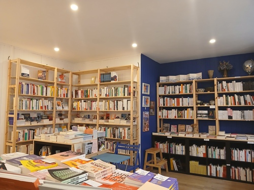 Photo de l'intérieur de la librairie, il y a un côté de murs blancs et un coté de murs bleus couverts de livres à chaque fois, et au centre des tables où sont exposés d'autres livres.