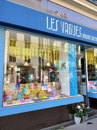 Devanture de la librairie, avec des livres exposés en vitrine et des dessins d'oiseaux prenant leur envol sur le verre. La façade est bleue avec écrit en blanc "Les Vagues Librairie queer"